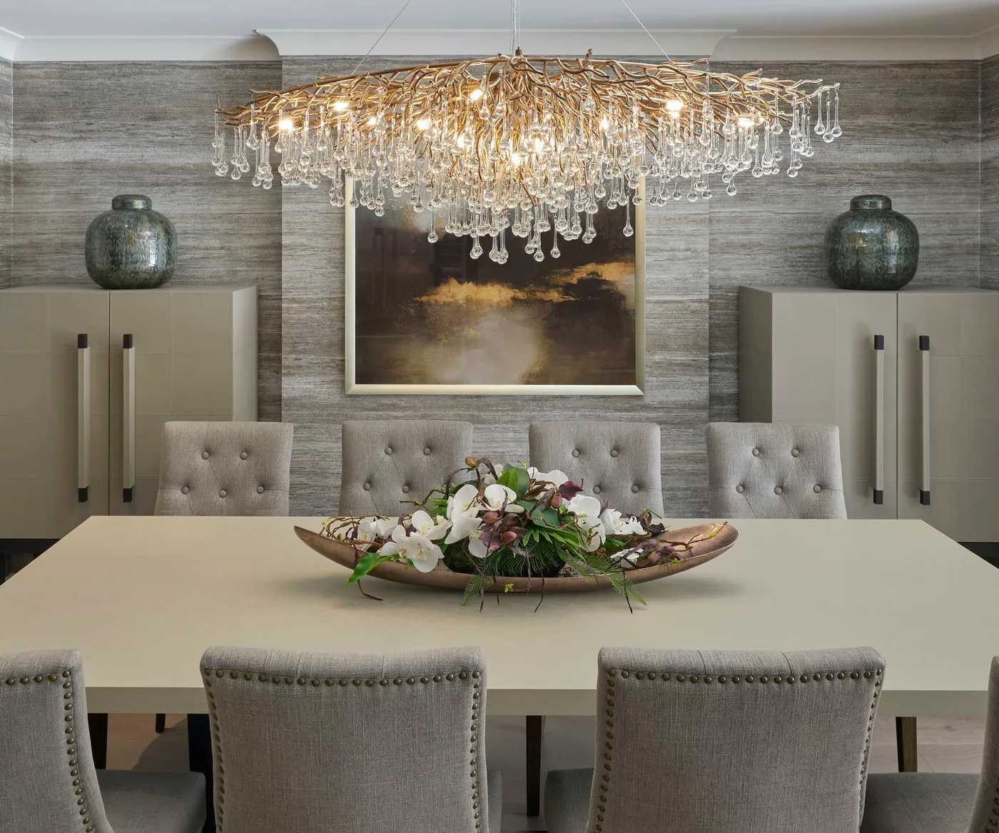 Dining room with a broad branching chandelier, pale table, tufted gray chairs, matching tall cupboards and green rounded vases