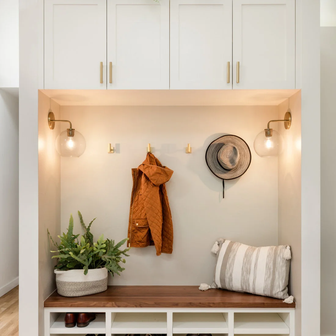 A wood bench with white shoe cubbies sits in a pale niche below coat hooks, overhead cupboards and two glass wall lights.