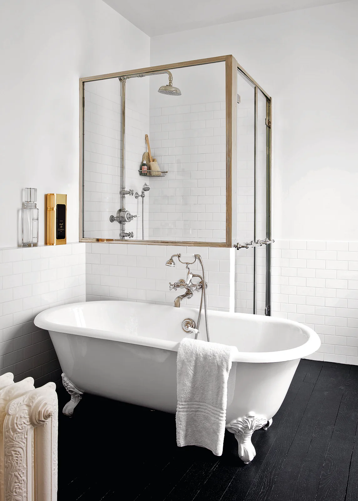 A white clawfoot tub sits on dark floorboards beside white subway tiles and a raised glazed shower with a warm-colored frame