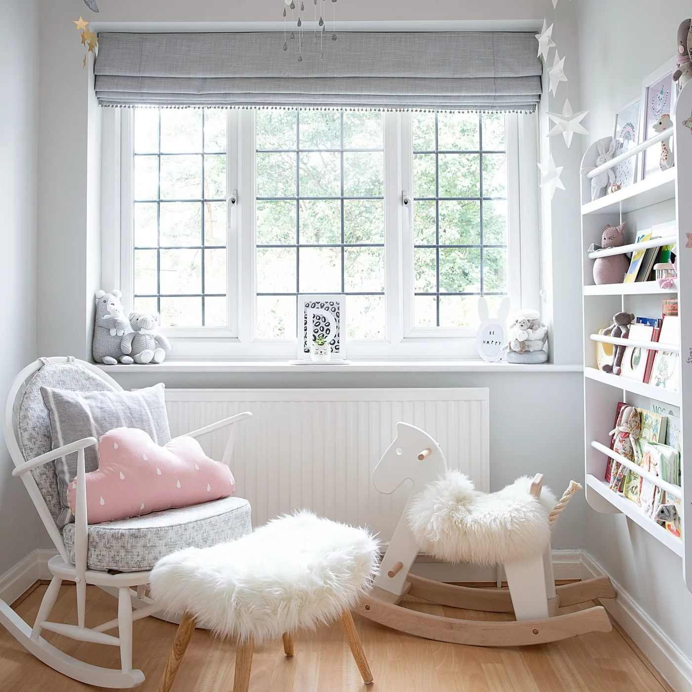Pale wooden chair with gray fabric and pink cloud cushion, small padded stool, rocking horse and open bookshelves by window.