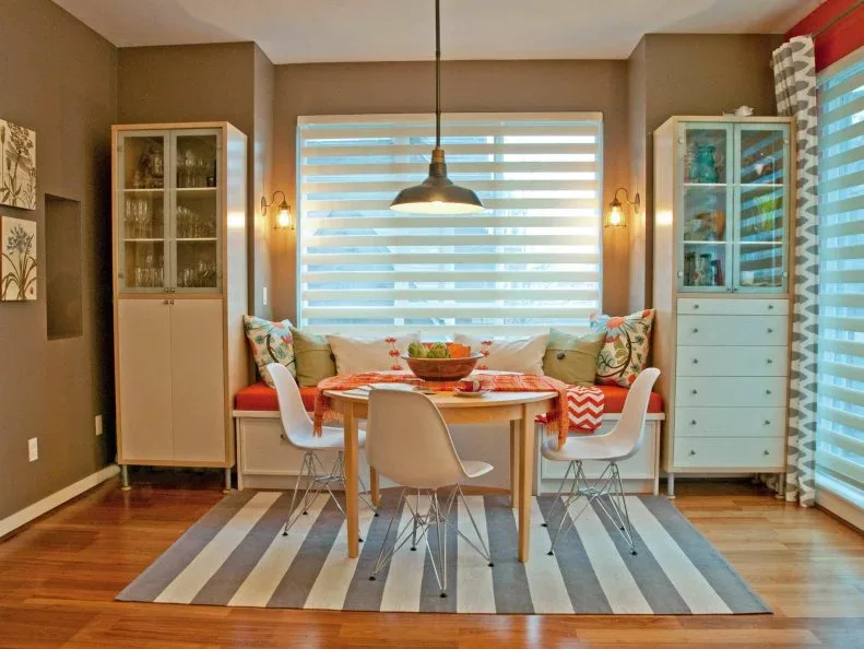 Dining nook with orange cushioned bench, white molded chairs, round wood table, striped rug and two pale display cupboards