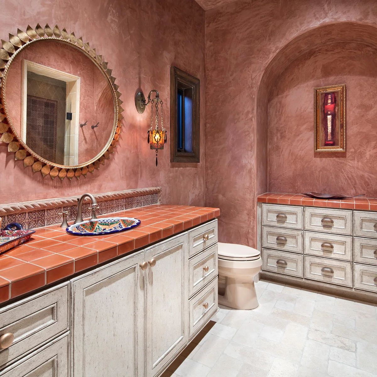 Warm pink bathroom with terracotta-colored tiled counters, patterned basin and pale distressed-looking cabinetry