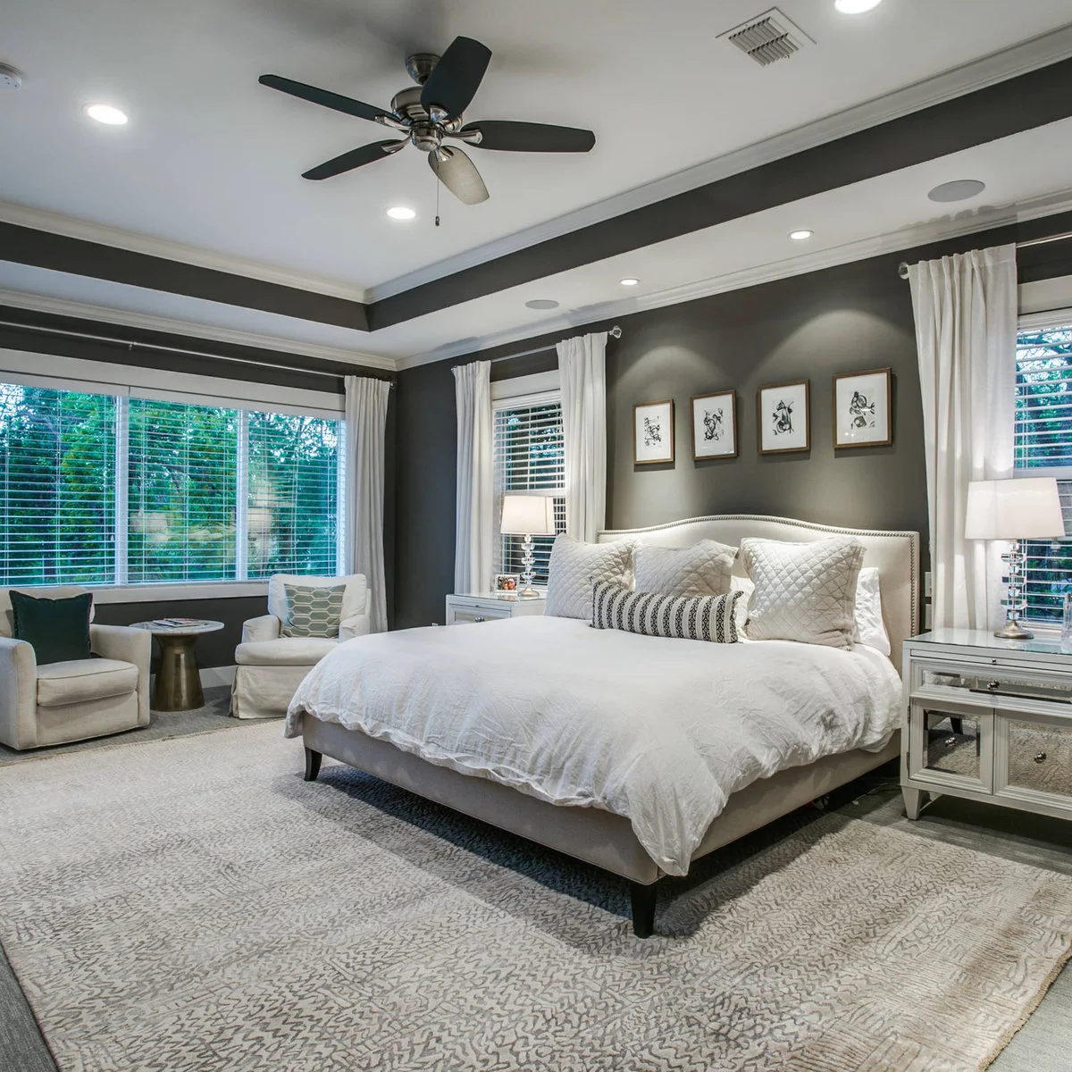 A pale bed and mirrored bedside chests in a dark gray room with two chairs beside the window and open floor around the bed.