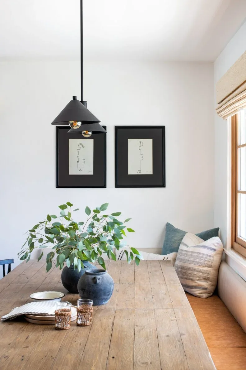 Country dining corner with wood table, cushioned bench beneath a window, black pendant and two framed pictures