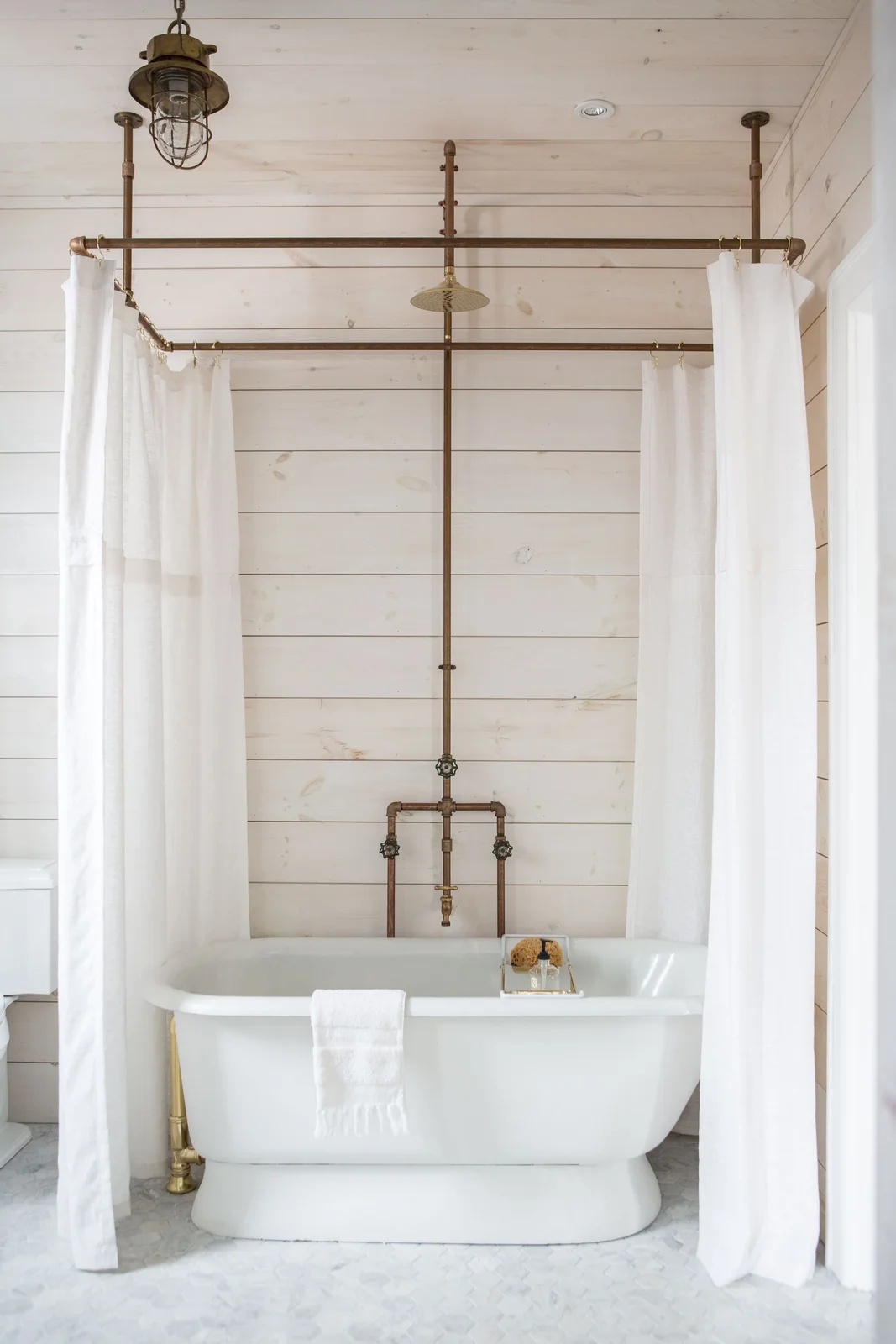 A white freestanding bath with exposed pipe shower fittings and white curtains against pale boards