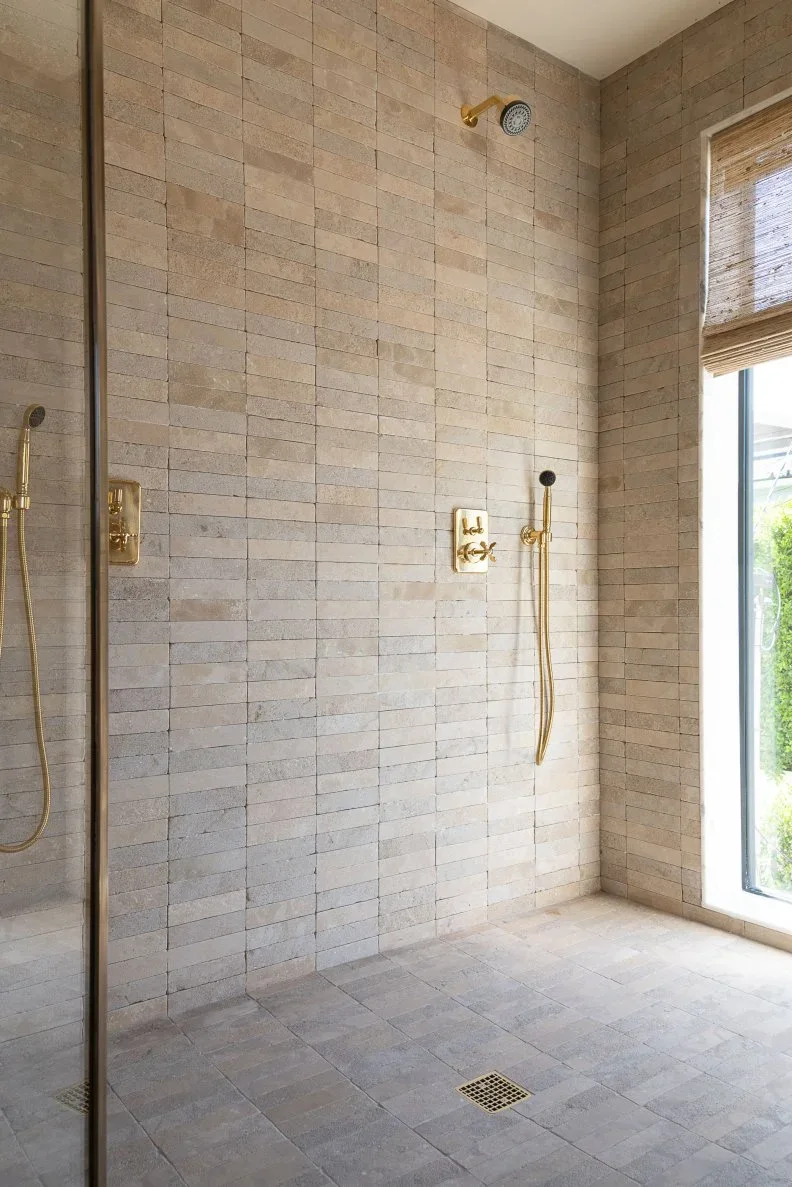 A shower lined with small pale rectangular tiles and warm metal fittings beside a tall window