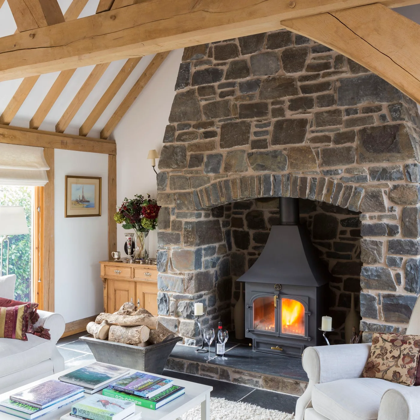 A tall dark stone chimney breast frames a stove beneath timber beams beside pale upholstered seating.