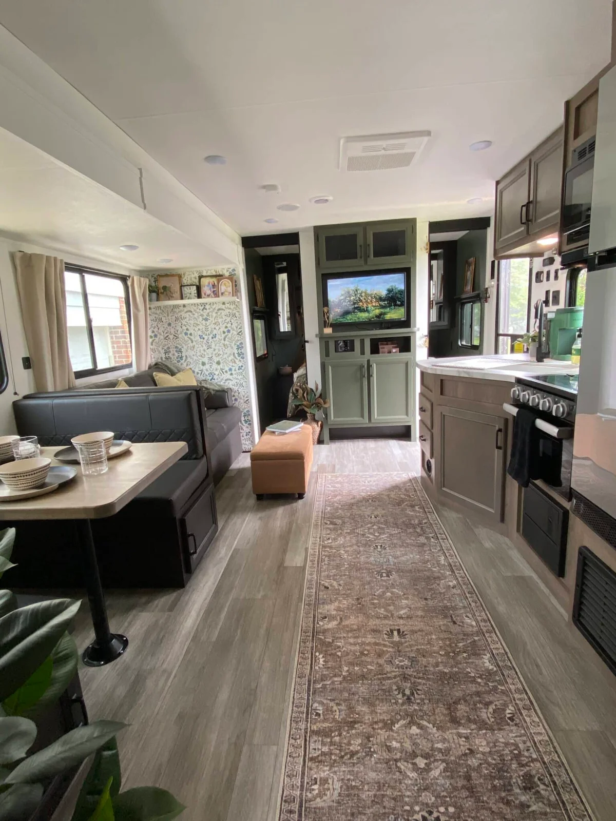 RV interior with gray cabinets, a patterned narrow runner, a dinette and a dark sofa with an orange ottoman.