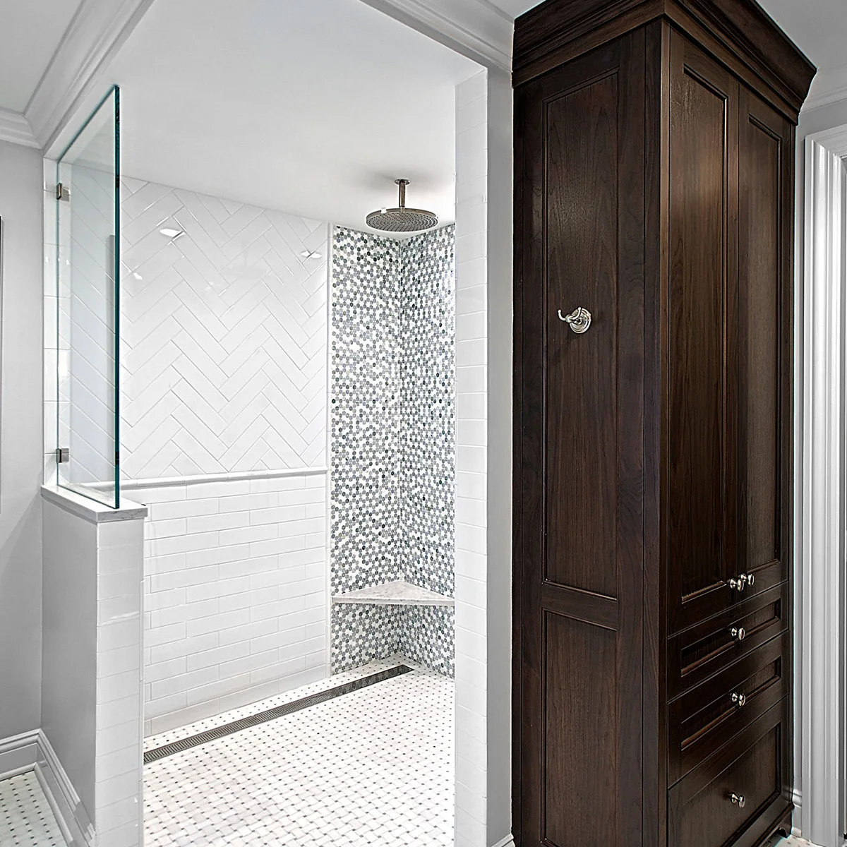 A white half wall and clear screen border a shower with herringbone tiles, a mosaic corner and a tall wood linen cabinet outside.