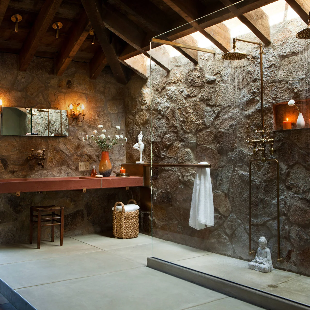A shower behind a clear glass screen with a rough wall, overhead daylight, exposed beams and warm-metal fittings beside a red-toned trough basin.