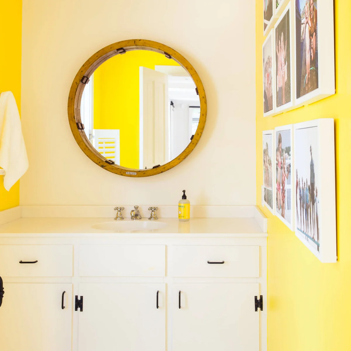 Large round wood-framed mirror above a white vanity beside yellow walls and a grid of family photographs.