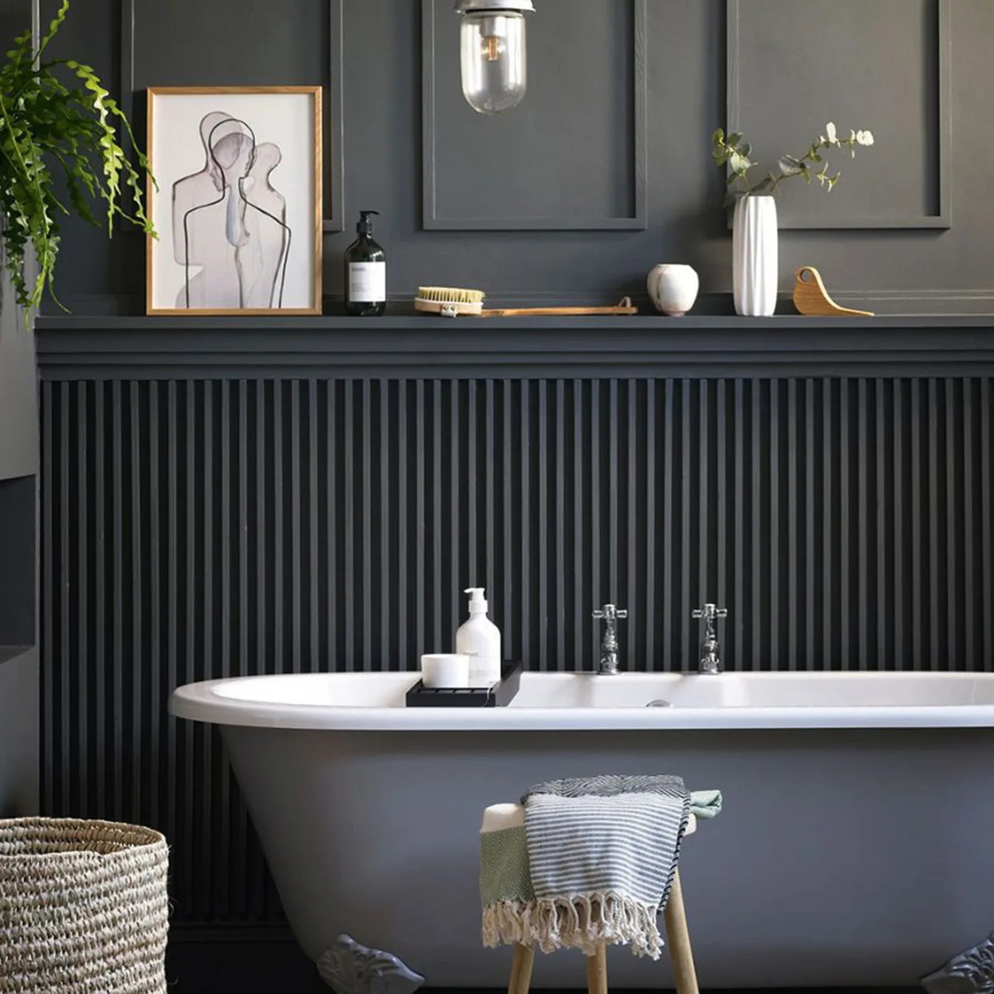 Gray bath below deep gray paneling with vertical ribbing, a narrow shelf, framed drawing, pale vases, trailing plant, glass pendant, and woven basket.