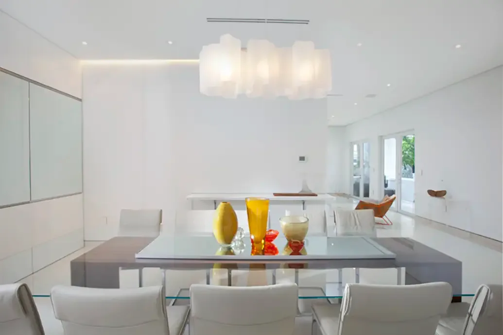 Clear glass dining table with white chairs and gray benches in a pale room with colorful vases