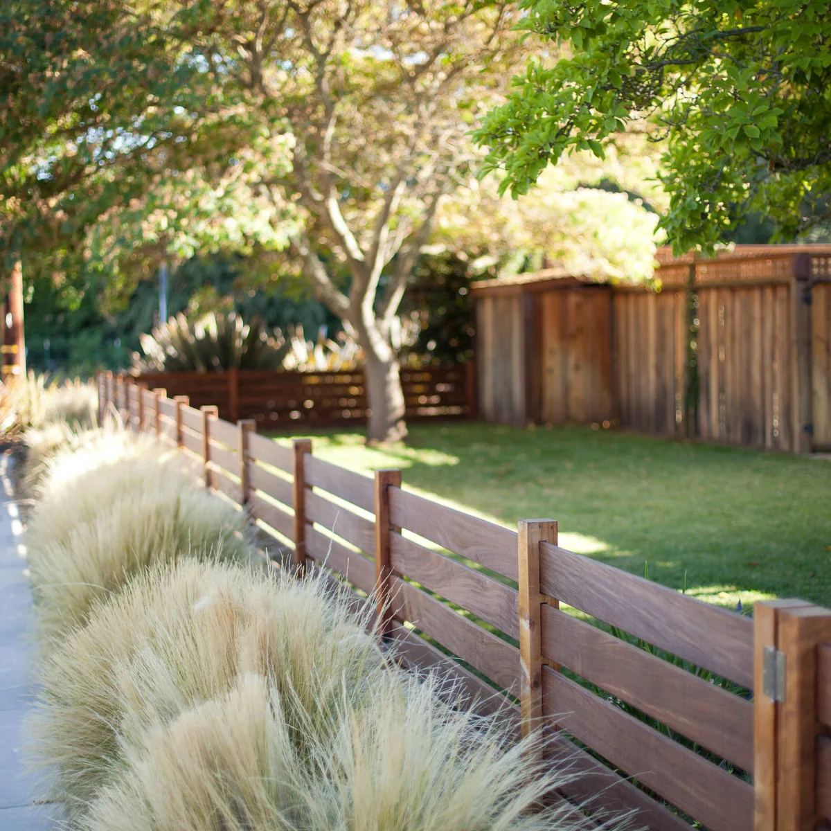 A low horizontal wood fence beside a sidewalk, with pale ornamental grasses outside and lawn behind.