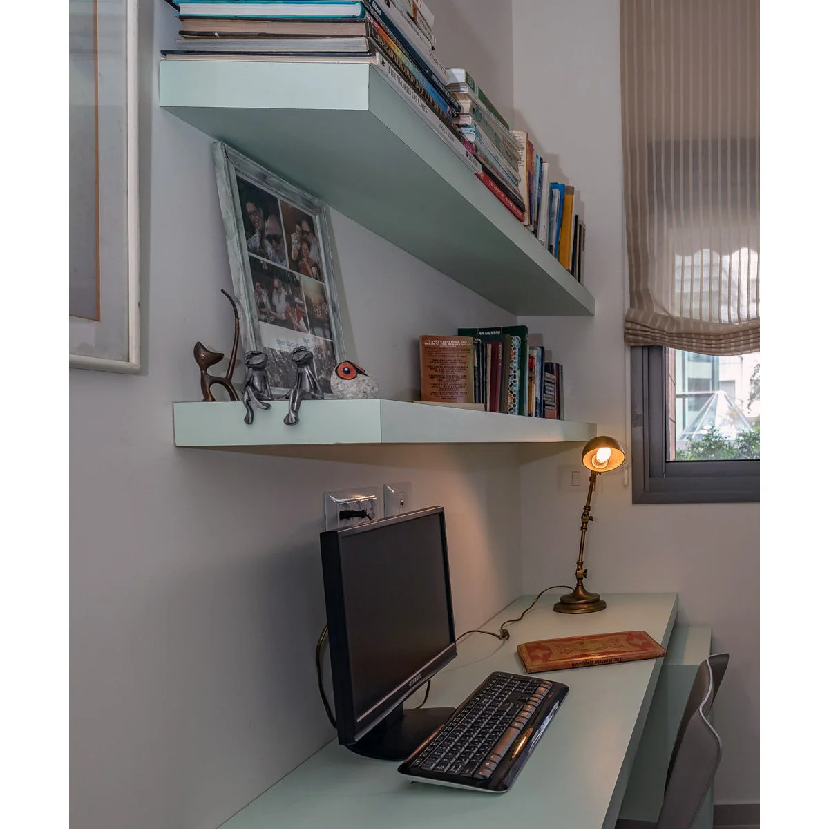 Compact office with pale desk, two wall shelves of books, small metal lamp and fabric window shade