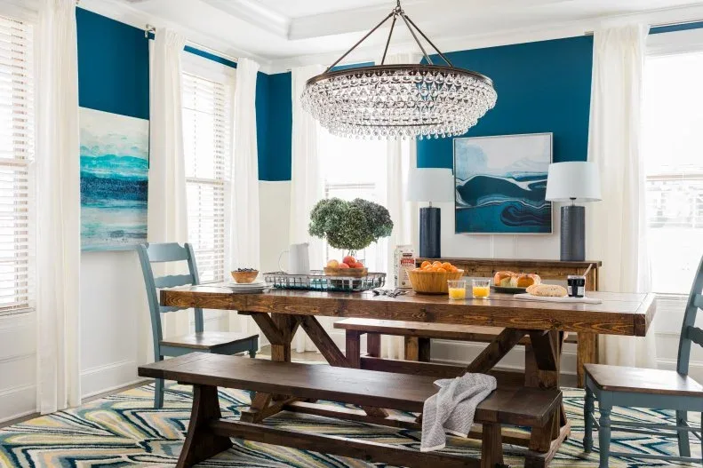 Dining room with high white paneling, deep blue upper walls, a wood table and bench beneath a broad chandelier