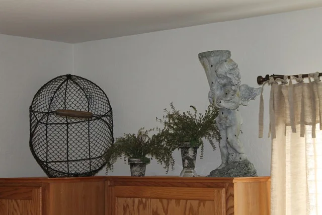 Large upright black wire basket, two small pots of greenery and a tall decorative vessel above wood kitchen cabinets.
