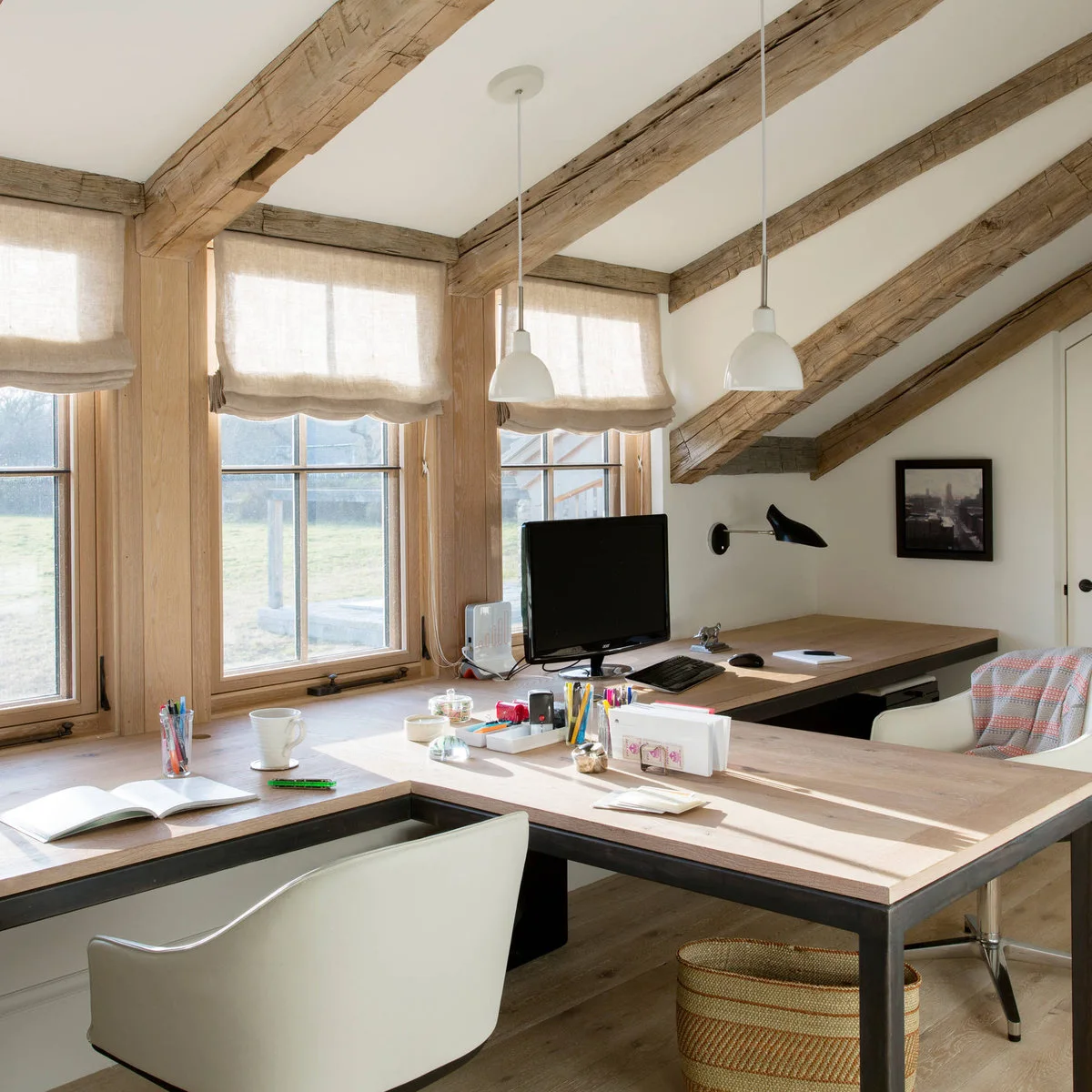 Wood desk below attic windows with a perpendicular peninsula, pale chairs and exposed ceiling beams