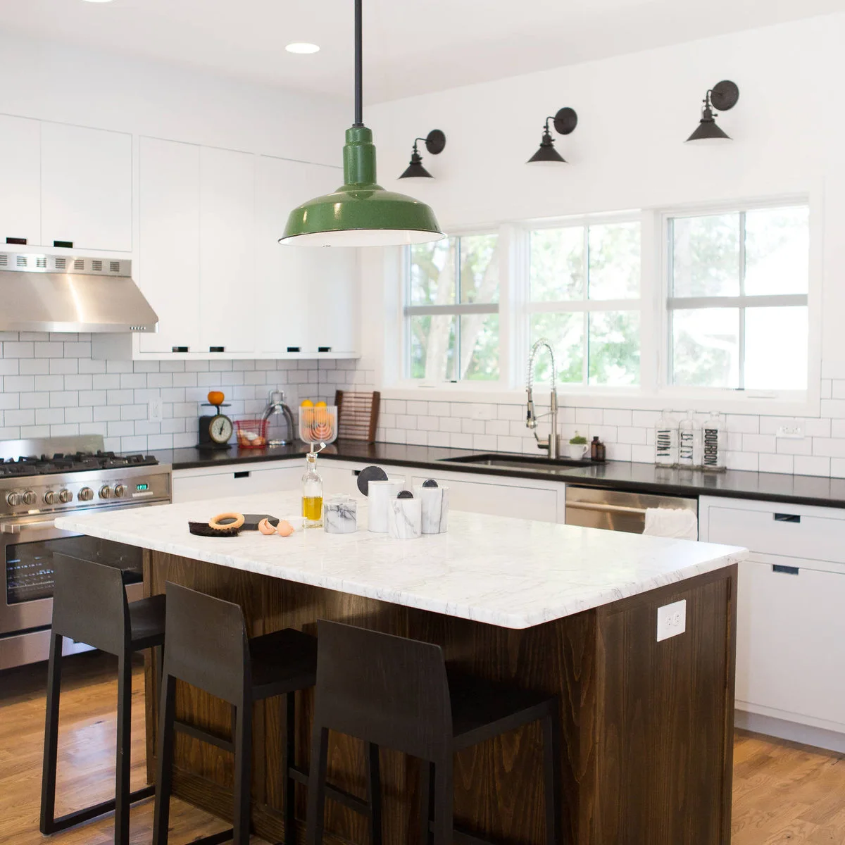 A green broad-shaded pendant hangs above a wood-sided island in a white kitchen with dark wall lights above the windows.