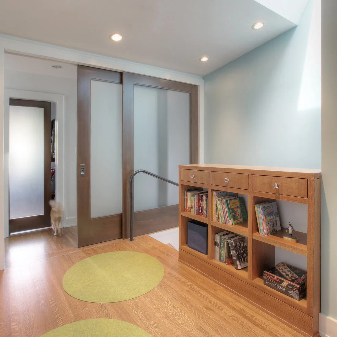 A wood cubby cabinet and frosted sliding doors sit beside pale blue walls and small round green rugs on a wood floor.