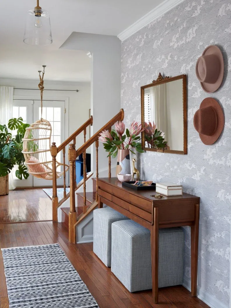Country console against floral wallpaper with flowers, a small tray and white box, two striped stools and hats on the nearby wall