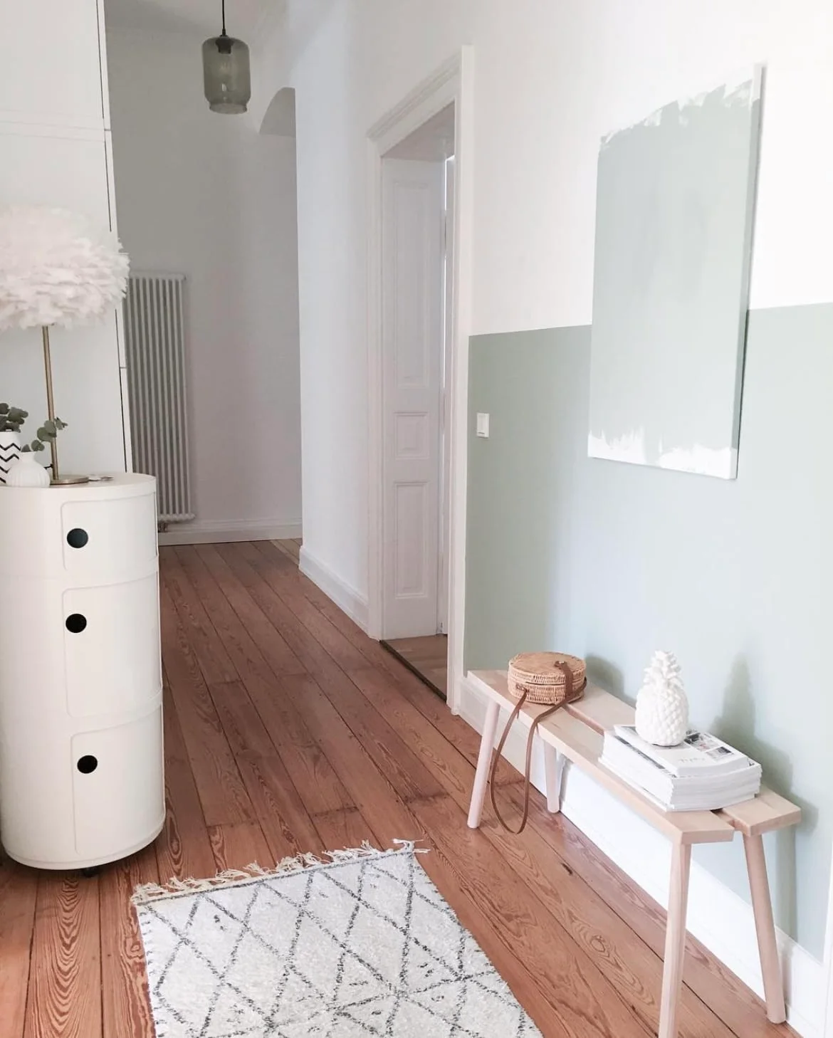 A small pale wooden bench stands against a pale green lower wall beneath light artwork beside a white storage unit and patterned rug