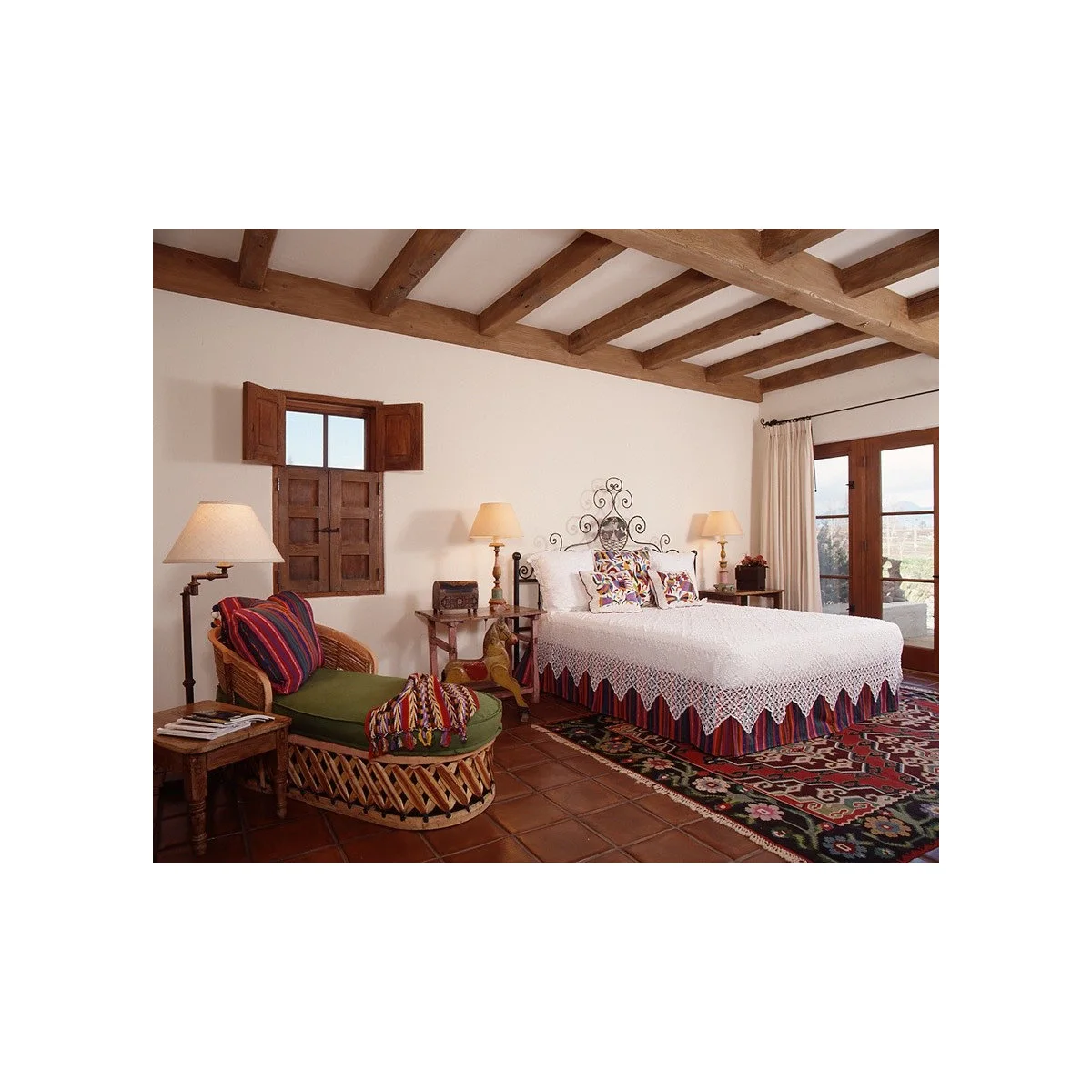 White textured bedcover with colorful cushions on a metal bed beside wood shutters, ceiling beams and a woven chair.