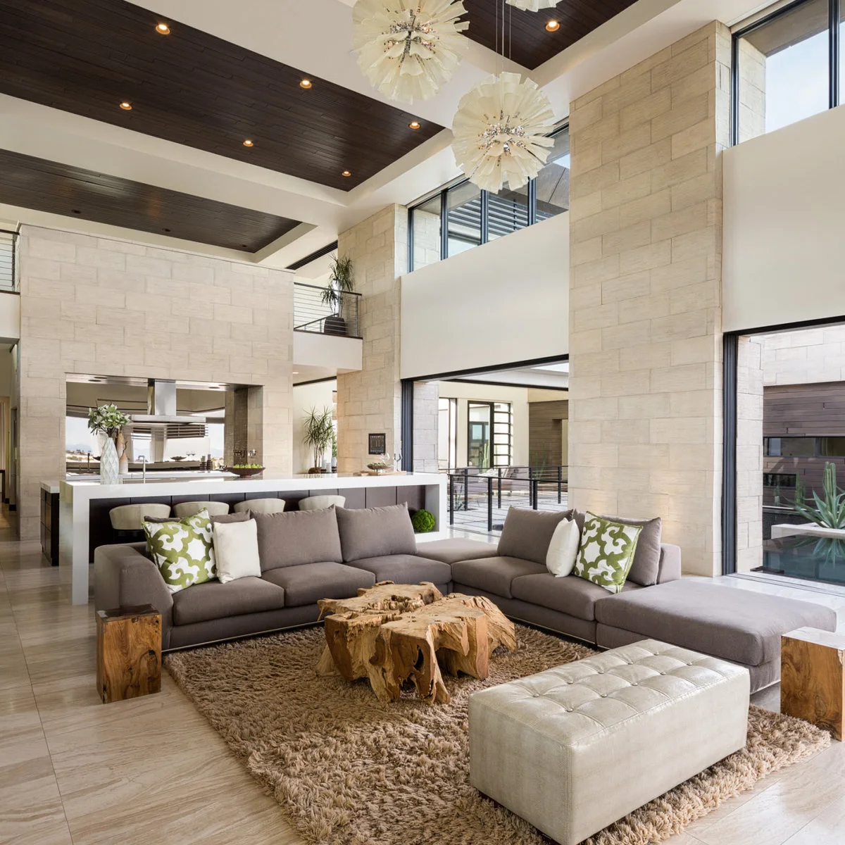 A high modern living room with a gray sectional, a thick brown rug, a sculptural wood coffee table and dark ceiling panels.