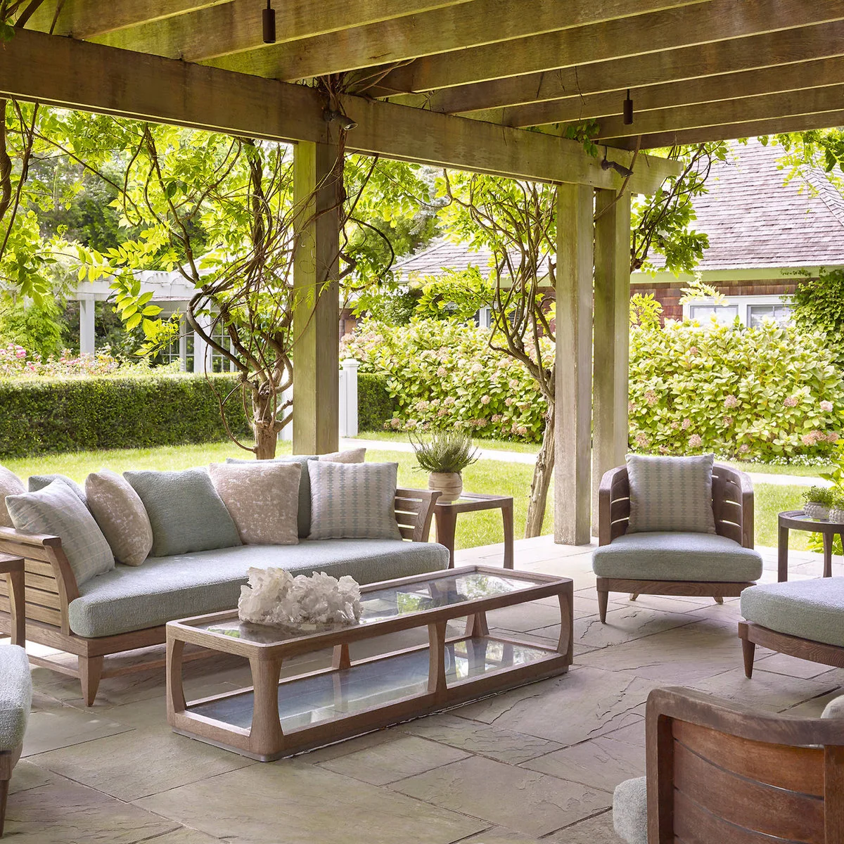 Climbing vines grow around an open wood pergola above pale gray lounge furniture and a glass-topped table.