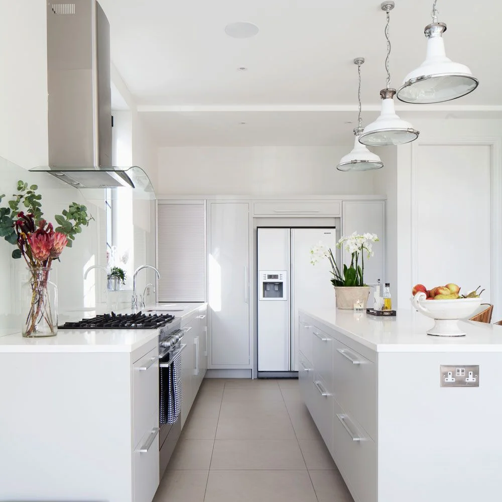 White framed tall cupboards surround a refrigerator opposite plain white island drawers beneath industrial-style pendants.