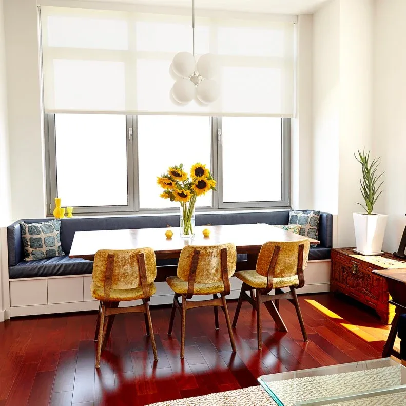 Dark upholstered straight banquette on a white paneled base beneath broad windows, with a wooden rectangular table, three mustard-colored chairs, sunflowers, and a white globe light.
