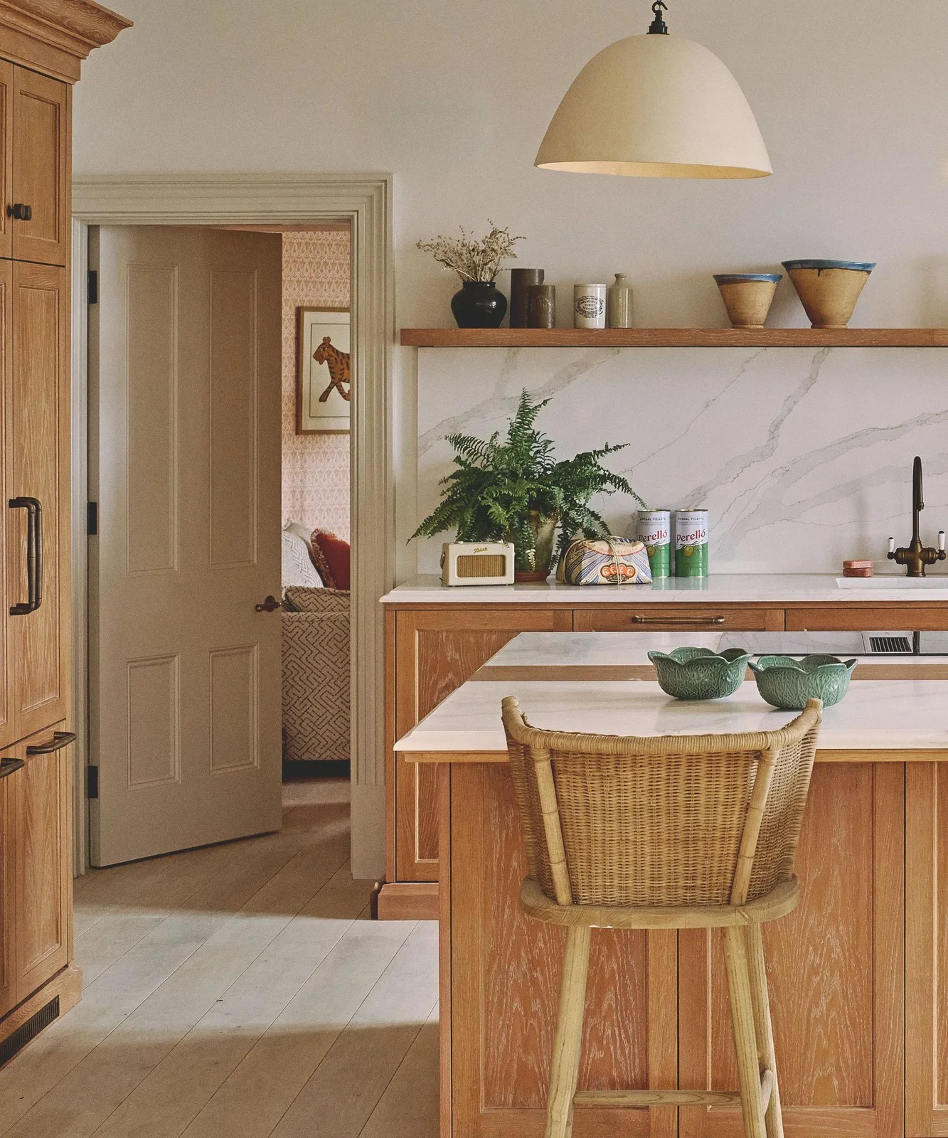 A woven backed stool beside timber cabinets, pale veined backsplash and open pottery shelf