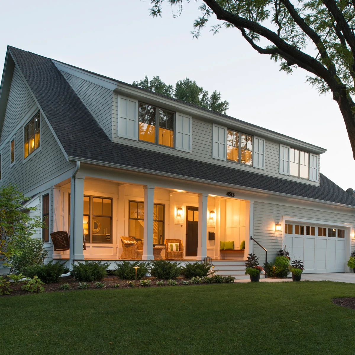 A pale Cape-inspired house has a wide shed dormer above a covered porch with square posts and woven chairs beside an attached garage.