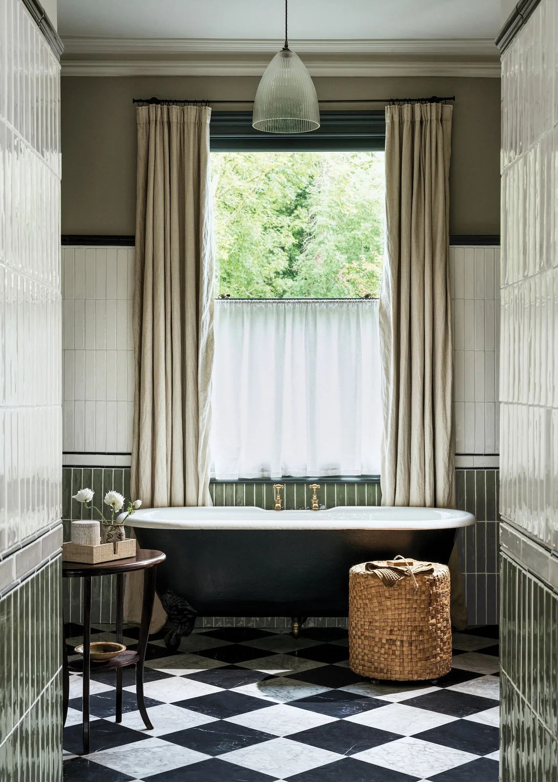 Glossy olive vertical tiles sit below white vertical tiles around a black freestanding bath, with a black-and-white checked floor.