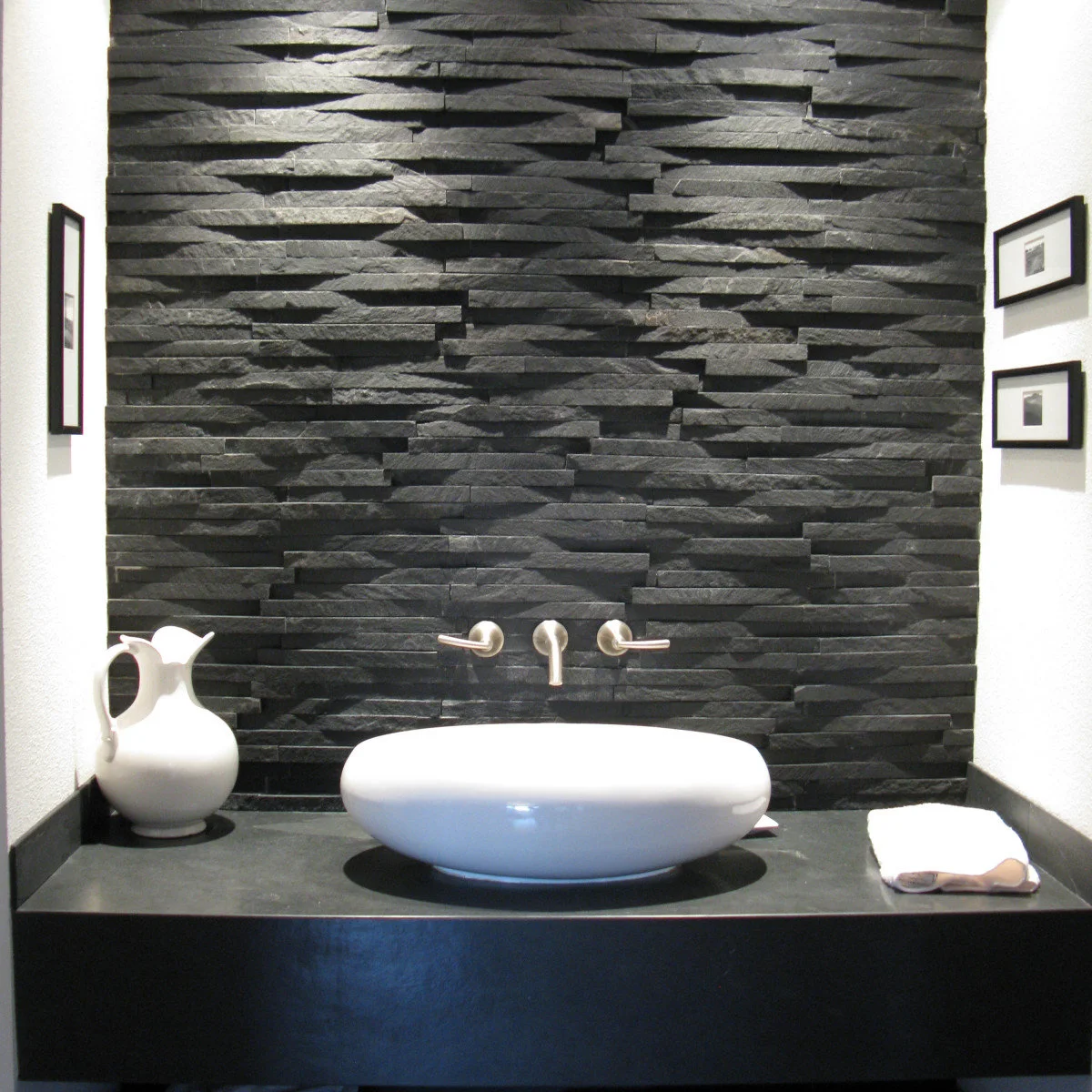 A smooth white oval vessel sink sits on a dark counter against a heavily textured dark wall with three wall-mounted faucet fittings.