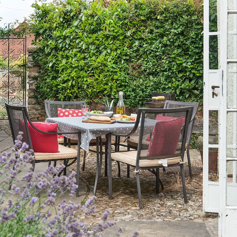 A dark dining table and chairs with red cushions sit beside a densely planted courtyard wall
