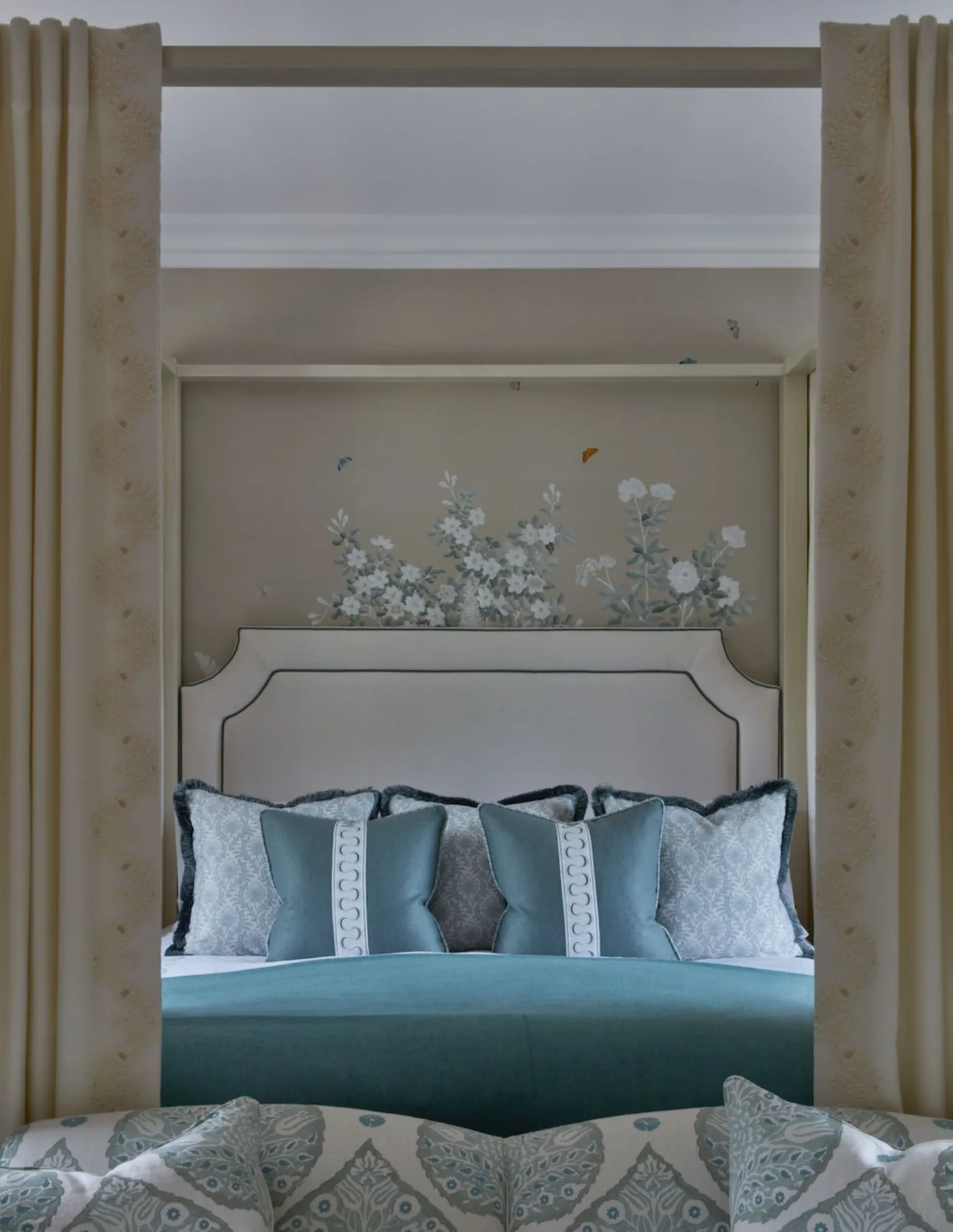 Traditional bed with a shaped pale headboard, curtains at either side, blue bedding and a small floral wall detail.
