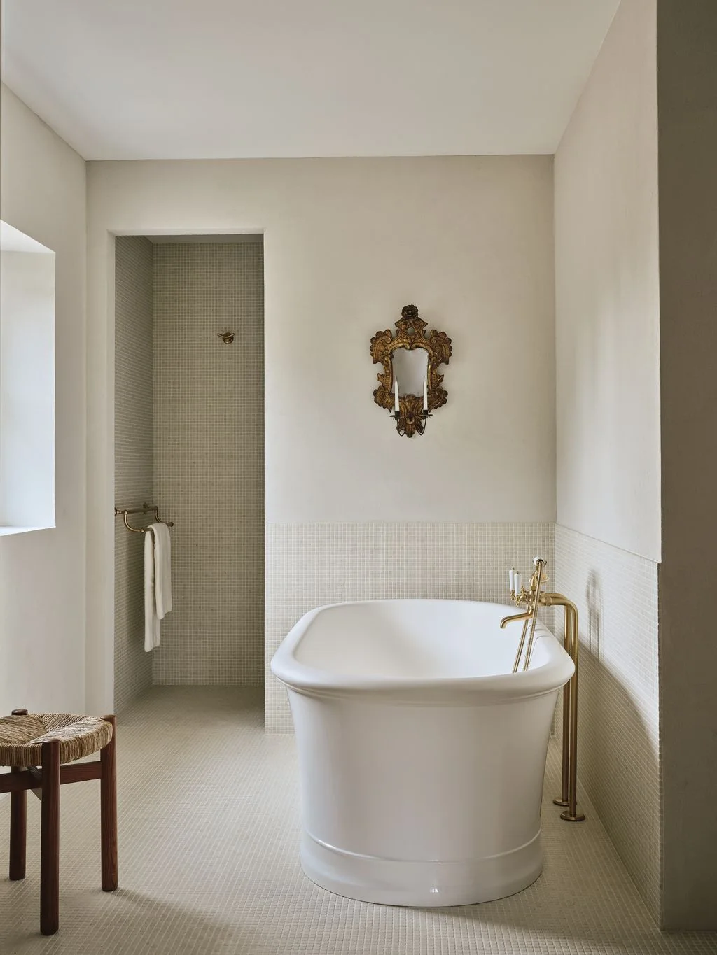 A tall open shower alcove lined with pale mosaic tiles sits beside a white freestanding bathtub and brass-colored floor faucet.