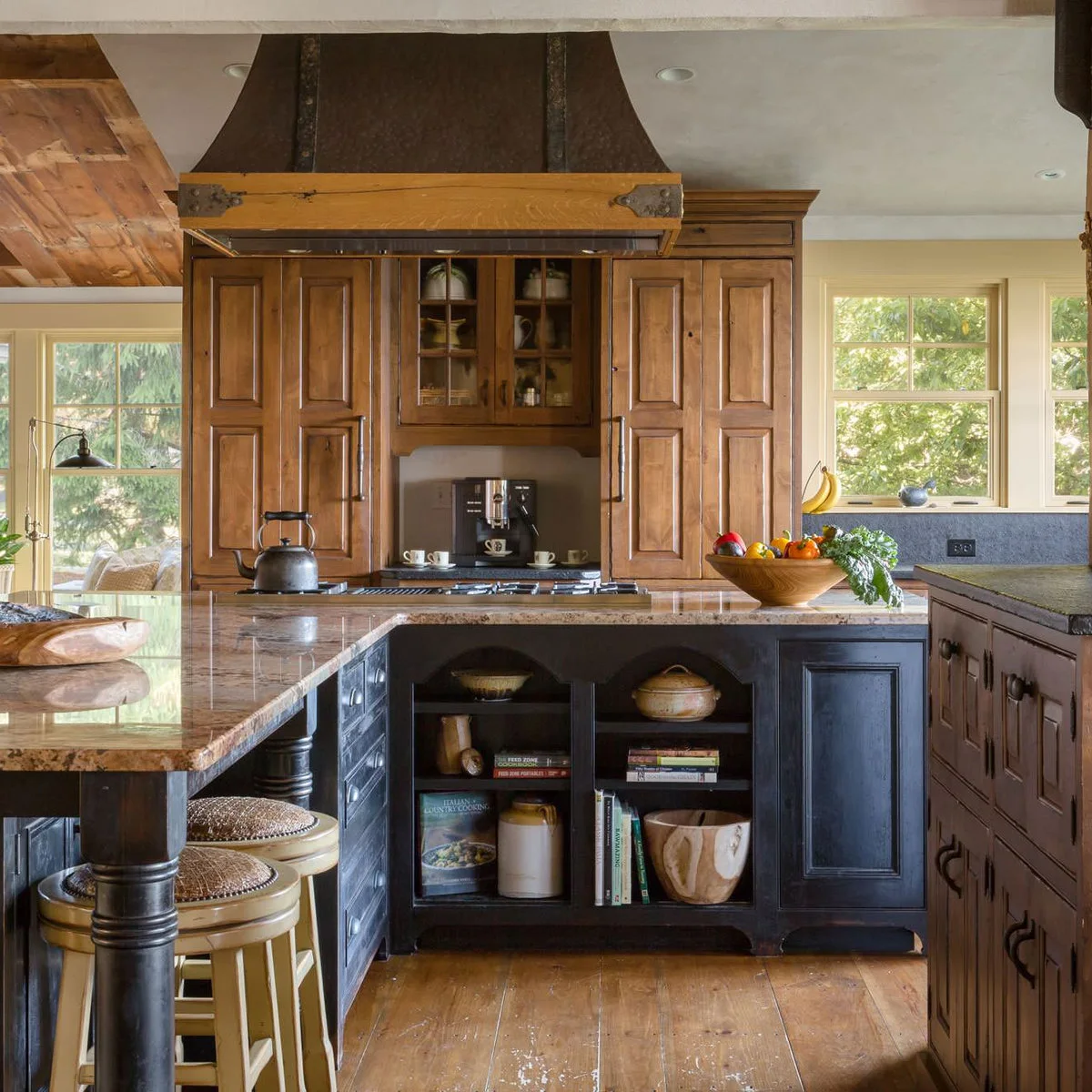 Warm wood cabinets and ceiling boards surround a dark island with a pale patterned counter and light stools.