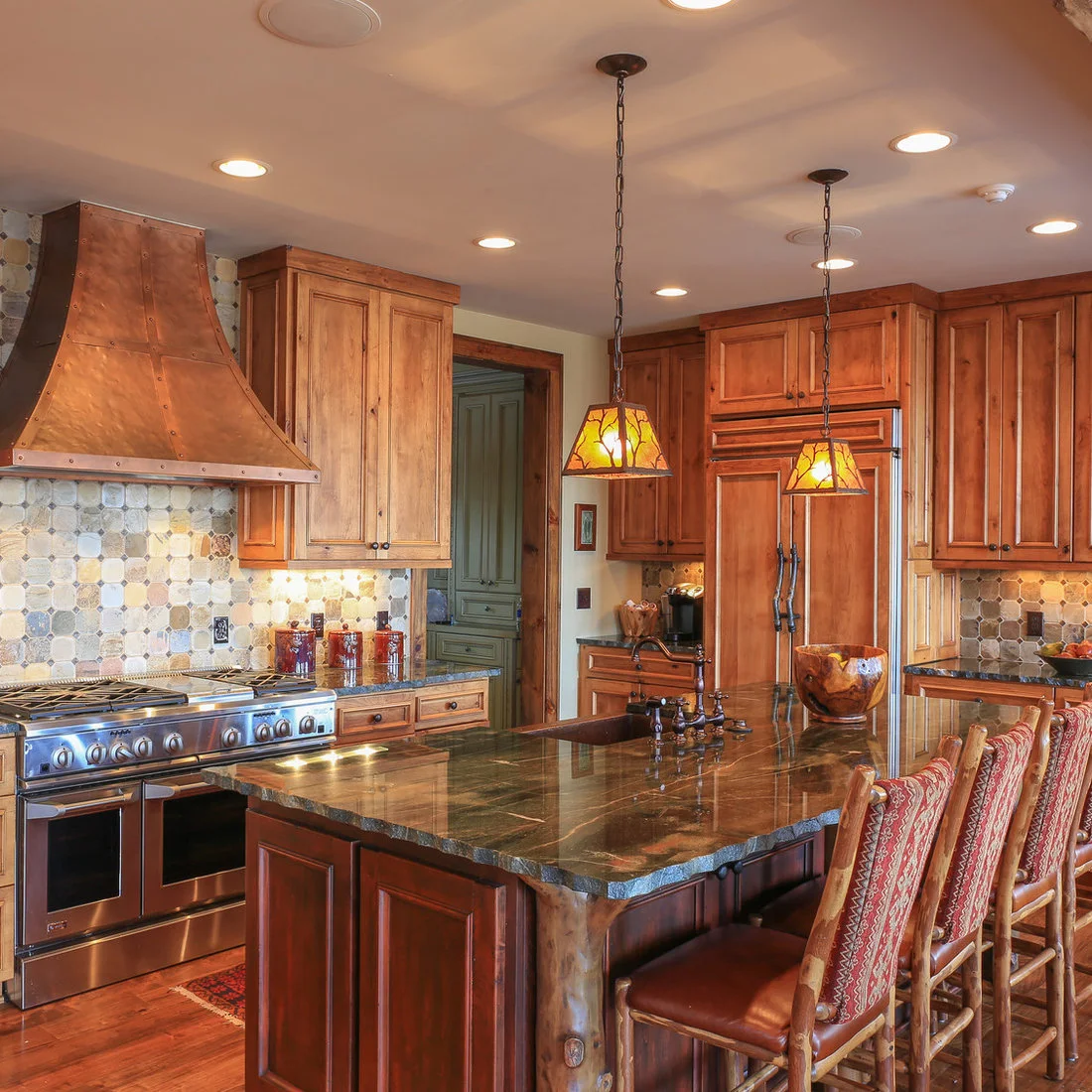 A warm metal range hood above patterned tiles beside wood cabinets and colored glass pendants