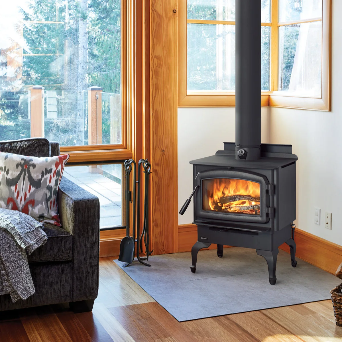 A black stove on a pale rectangular hearth sits beside timber-framed windows, with a dark chair and a metal tool stand nearby.