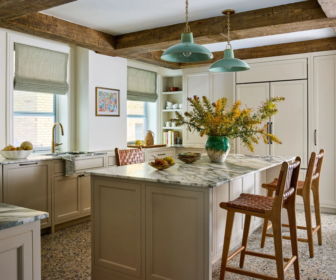 An off-white kitchen has exposed ceiling beams, green dome pendants, striped blinds, a veined island top and brown woven-back stools.