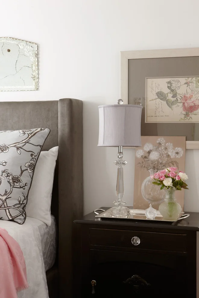 Dark bedside cabinet with a clear lamp, pale flower arrangement and tray beside a gray headboard