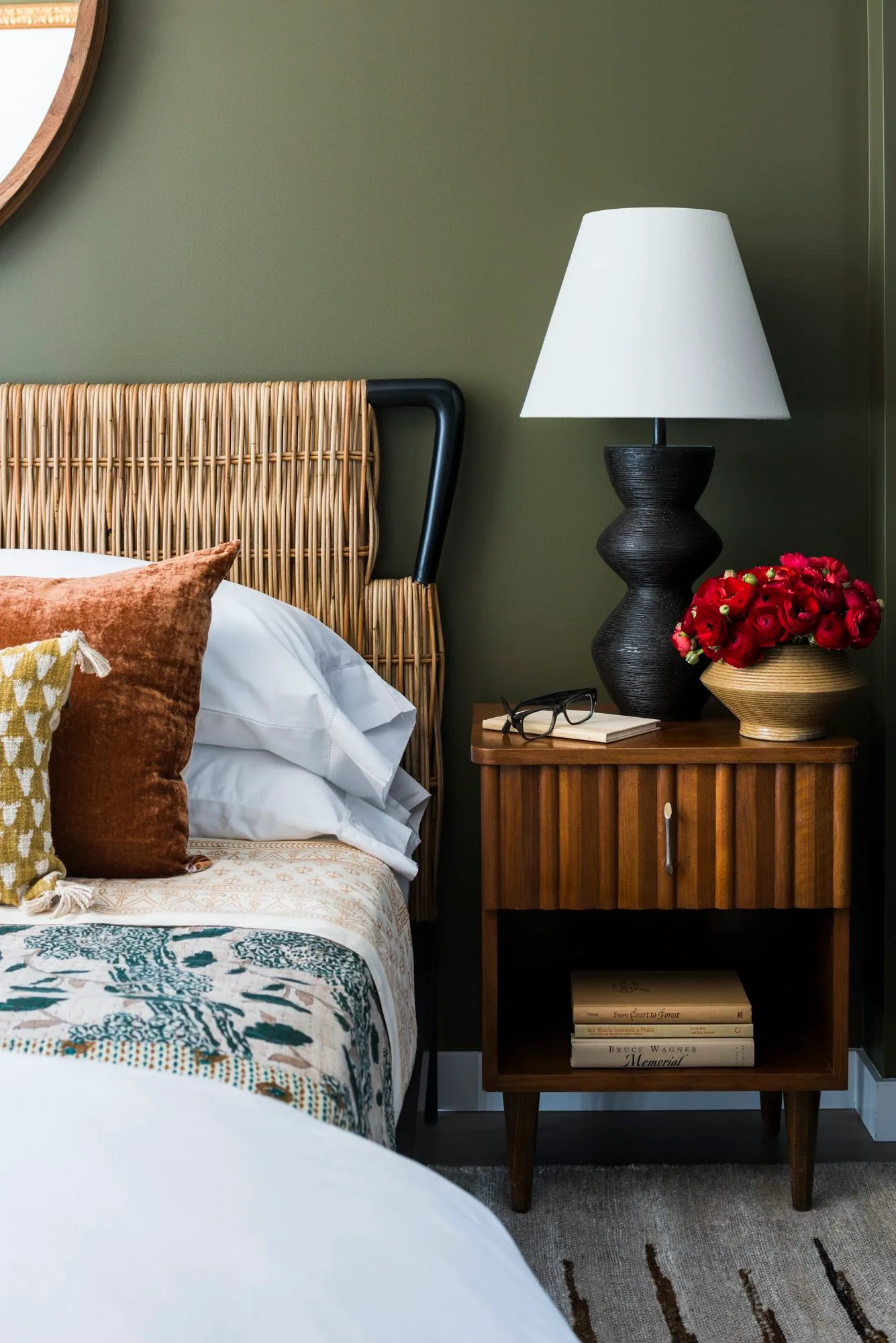 Dark olive bedroom with a woven headboard in a black frame, rust cushion, white bedding and a fluted wood bedside cabinet