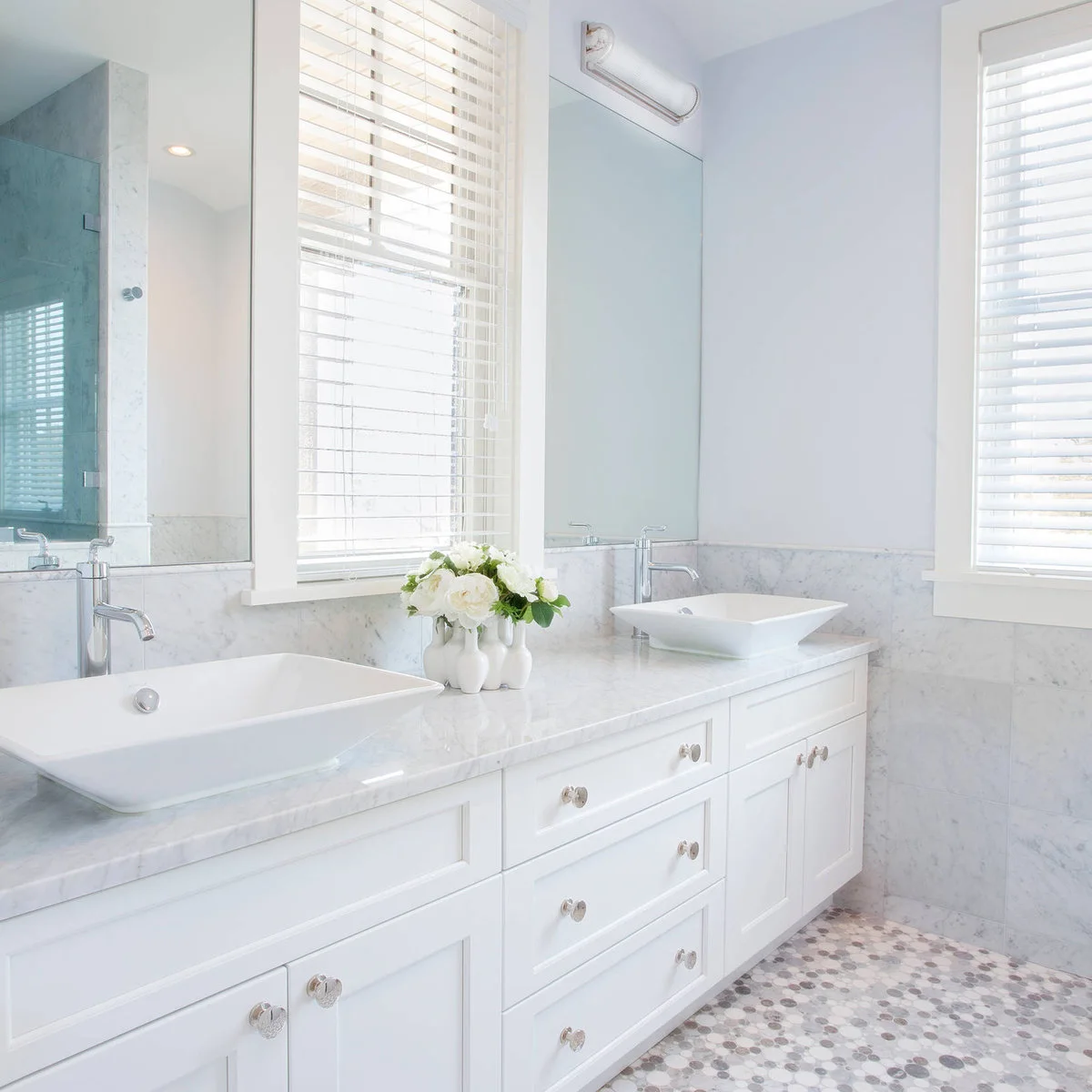Two white flared rectangular basins sit on a pale veined counter above white drawers, with a window between broad mirrors.