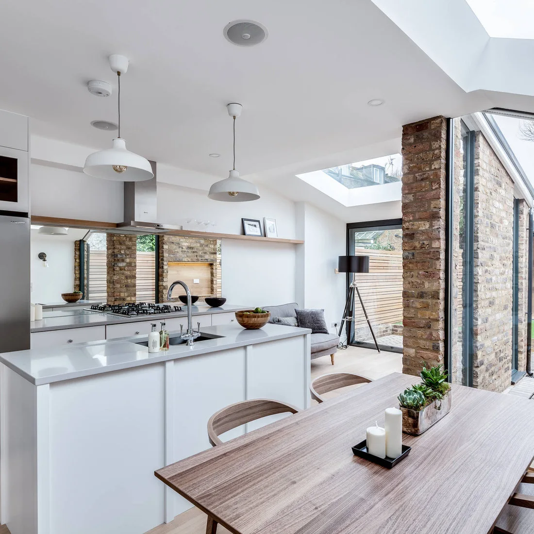 A long wood table and curved wood chairs stand beside a white kitchen island, rooflights and exposed brick piers.