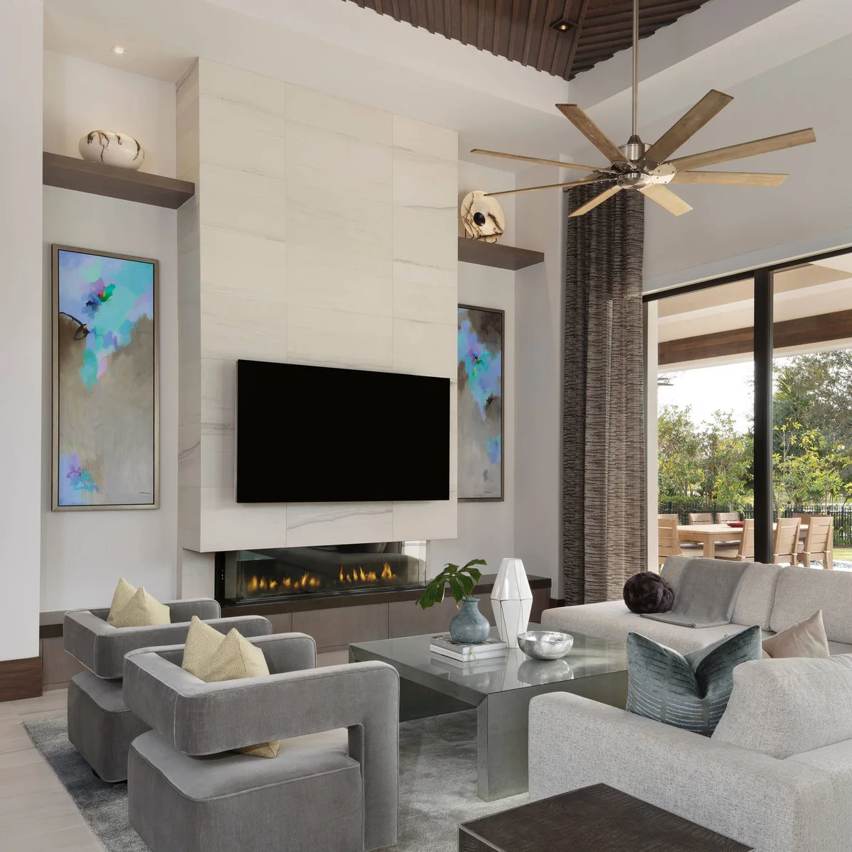 A tall living room with a pale fireplace surround, vertical paintings, gray seating, dark curtains and a large ceiling fan.