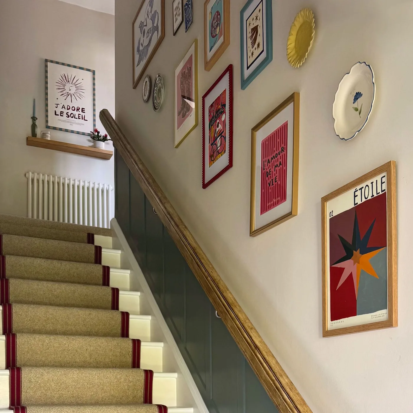 Entry staircase with neutral runner edged in red, wood handrail, framed colorful pictures and decorative plates along the stair wall