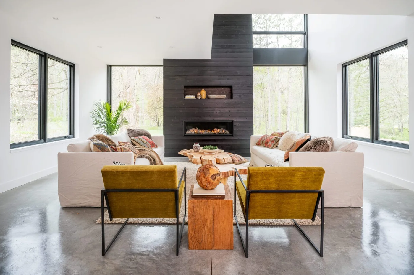 Two mustard-colored chairs face pale sofas and small wood tables around a tall dark fireplace wall beside broad windows and a gray floor