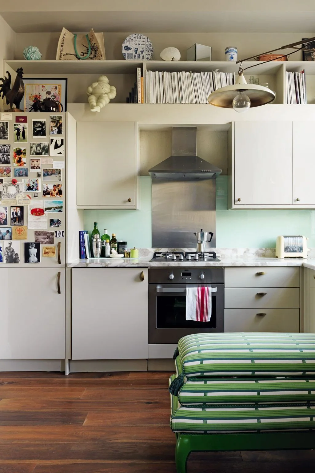 Plain pale kitchen cabinets surround a compact cooking run beneath shelves of books and objects beside a refrigerator covered with photographs.